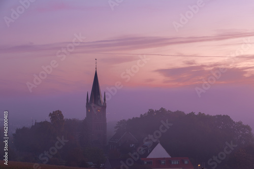 A typical colorful Autumn sunrise in Maastricht with the landscape covered with a layer of fog, leaving only silhouettes visible in the distance, like this tower of a church on the hillside.