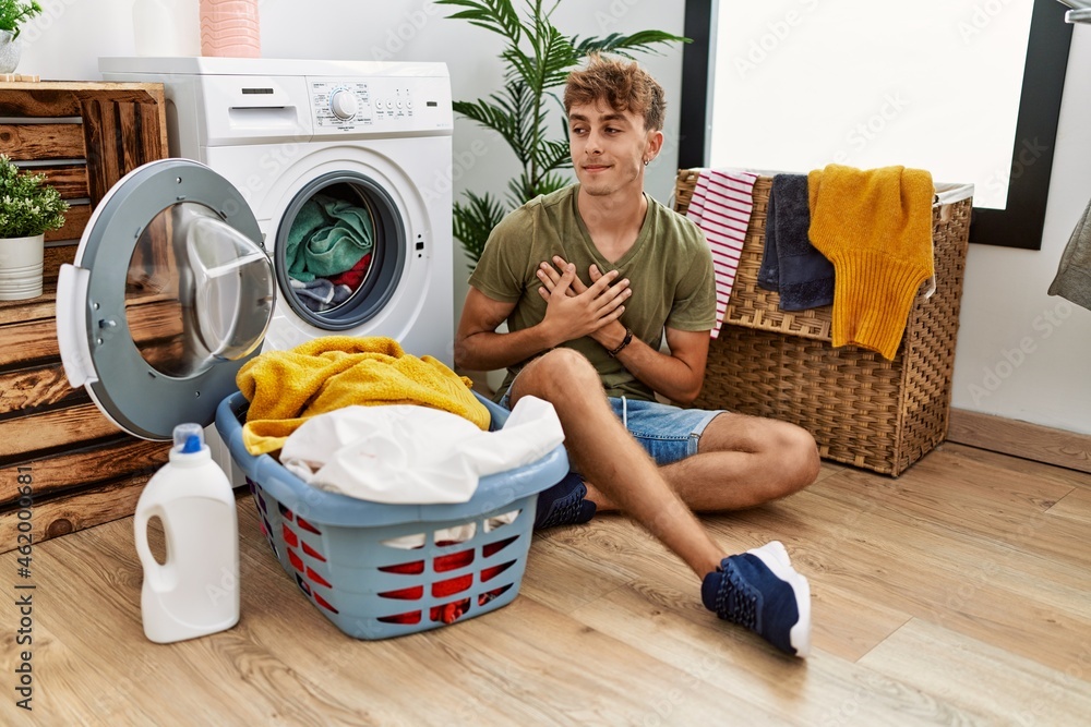 Young caucasian man putting dirty laundry into washing machine smiling with hands on chest with closed eyes and grateful gesture on face. health concept. Young caucasian man putting dirty laundry into washing machine smiling with hands on chest with closed eyes and grateful gesture on face. health concept.