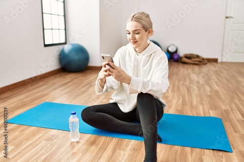 Young blonde girl relaxed using smartphone sitting on the floor at sport center.