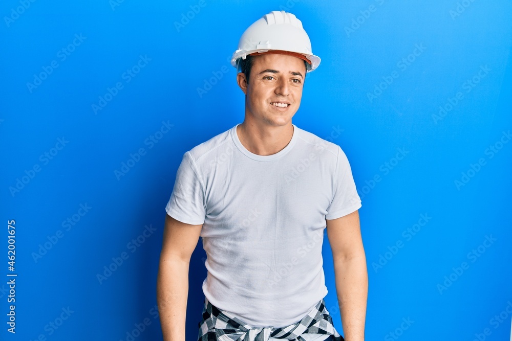 Handsome young man wearing builder uniform and hardhat looking away to side with smile on face, natural expression. laughing confident.