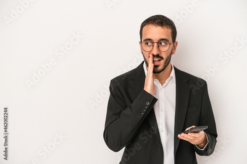 Young business man holding a mobile phone isolated on white background is saying a secret hot braking news and looking aside