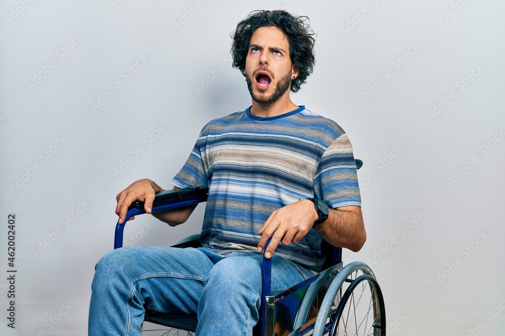 Handsome hispanic man sitting on wheelchair angry and mad screaming frustrated and furious, shouting with anger. rage and aggressive concept. Handsome hispanic man sitting on wheelchair angry and mad screaming frustrated and furious, shouting with anger. rage and aggressive concept.