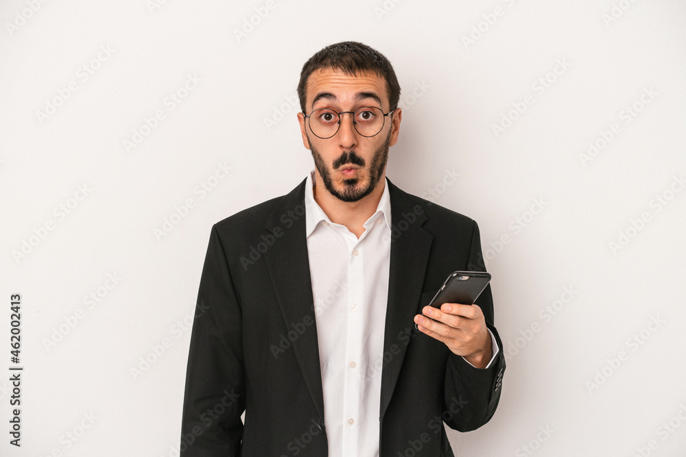 Young business man holding a mobile phone isolated on white background shrugs shoulders and open eyes confused. Young business man holding a mobile phone isolated on white background shrugs shoulders and open eyes confused.