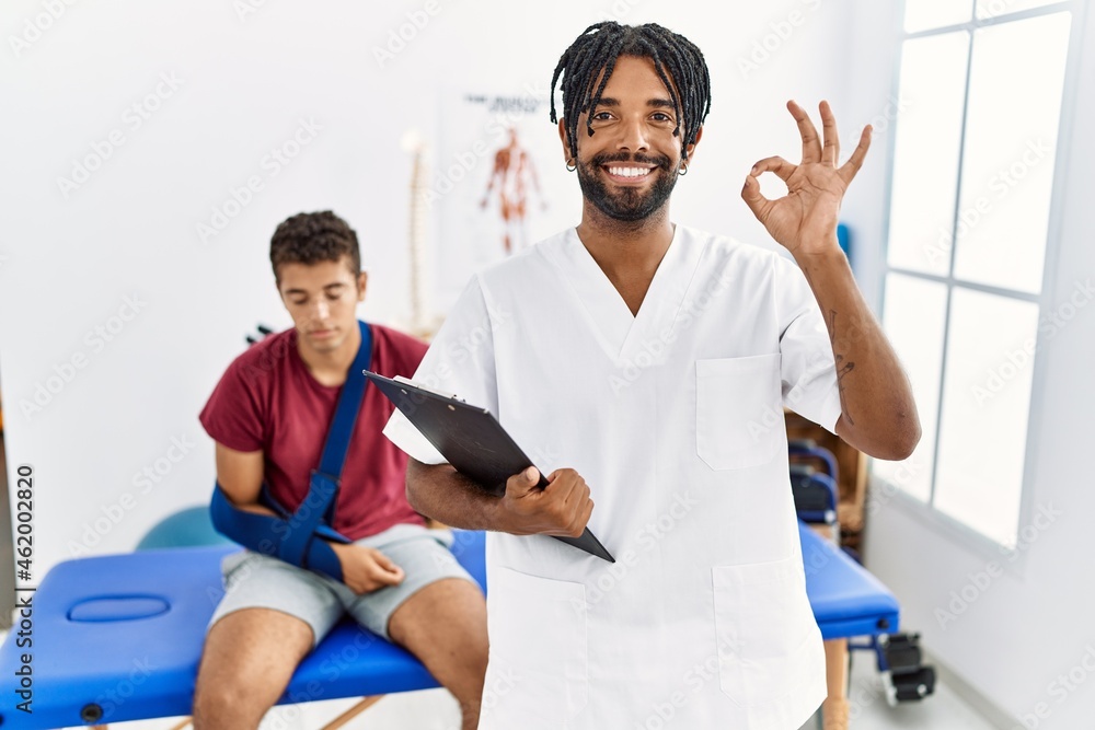 Young hispanic man working at pain recovery clinic with a man with broken arm smiling positive doing ok sign with hand and fingers. successful expression. Young hispanic man working at pain recovery clinic with a man with broken arm smiling positive doing ok sign with hand and fingers. successful expression.