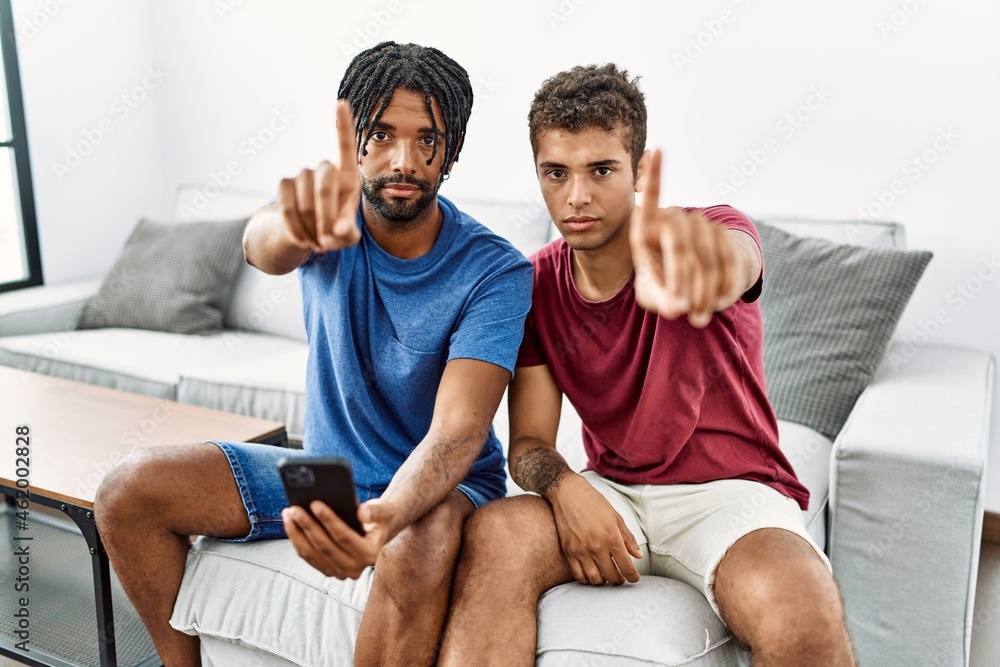 Obraz premium Young hispanic men using smartphone sitting on the sofa at home pointing with finger up and angry expression, showing no gesture