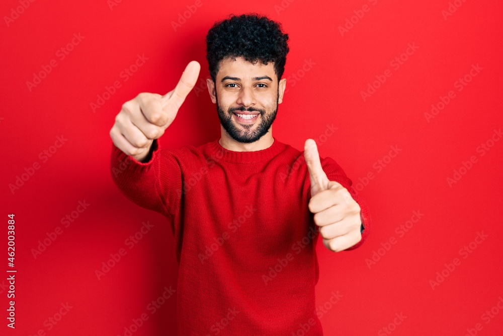 Young arab man with beard wearing casual red sweater approving doing positive gesture with hand, thumbs up smiling and happy for success. winner gesture. Young arab man with beard wearing casual red sweater approving doing positive gesture with hand, thumbs up smiling and happy for success. winner gesture.