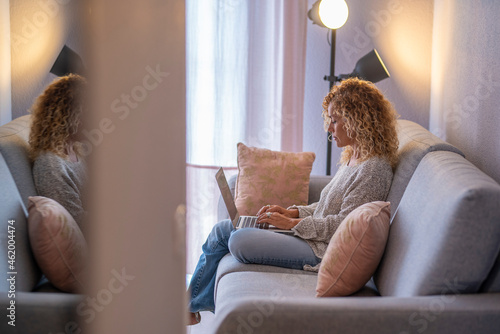 Young woman working on laptop sitting on sofa in living room at home. Caucasian businesswoman in casuals working from home. Caucasian woman typing on laptop in living room.