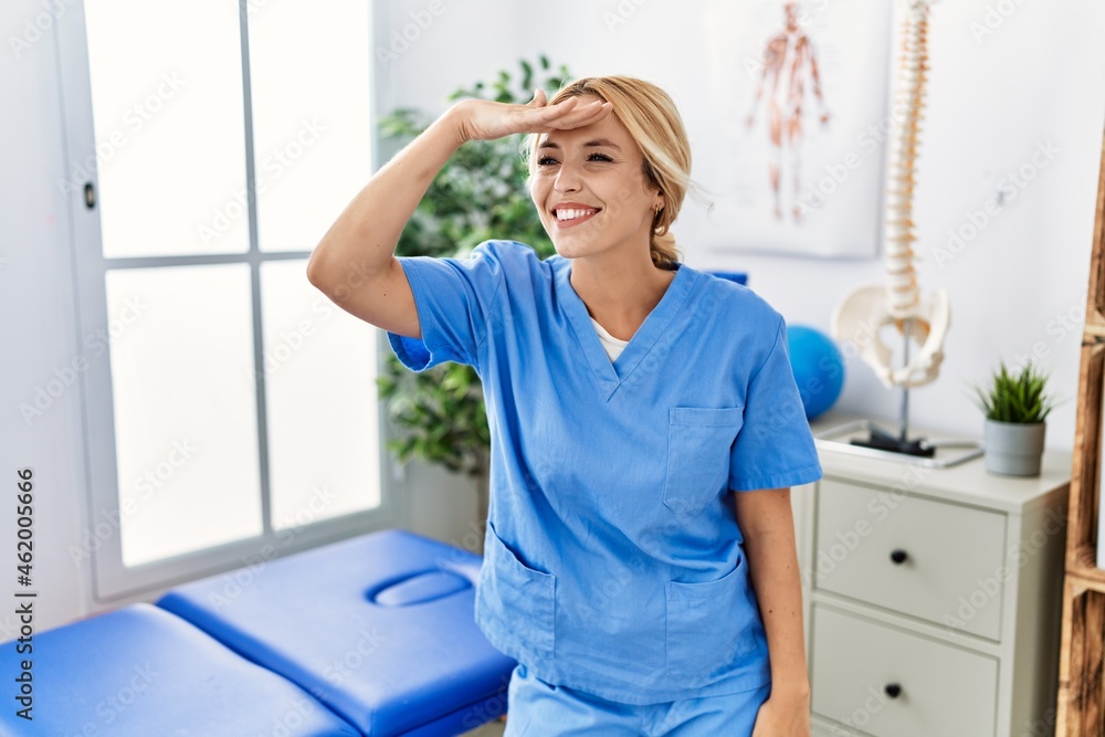 Fototapeta premium Beautiful blonde physiotherapist woman working at pain recovery clinic very happy and smiling looking far away with hand over head. searching concept.