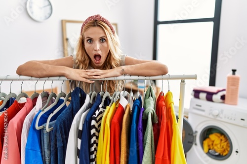 Beautiful blonde woman at laundry room with clean clothes afraid and shocked with surprise and amazed expression, fear and excited face.