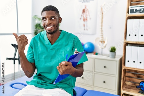 Young african american man working at pain recovery clinic smiling with happy face looking and pointing to the side with thumb up.