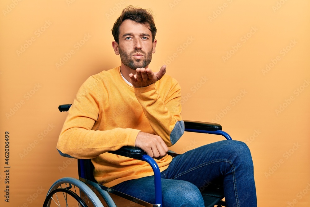 Handsome man with beard sitting on wheelchair looking at the camera blowing a kiss with hand on air being lovely and sexy. love expression. Handsome man with beard sitting on wheelchair looking at the camera blowing a kiss with hand on air being lovely and sexy. love expression.