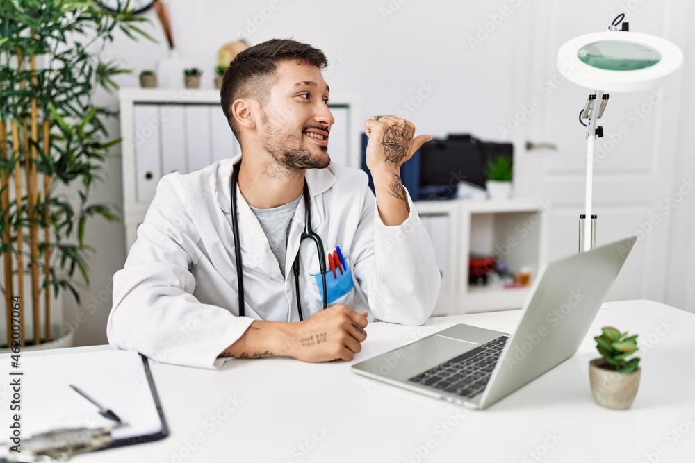Young doctor working at the clinic using computer laptop smiling with happy face looking and pointing to the side with thumb up. Young doctor working at the clinic using computer laptop smiling with happy face looking and pointing to the side with thumb up.