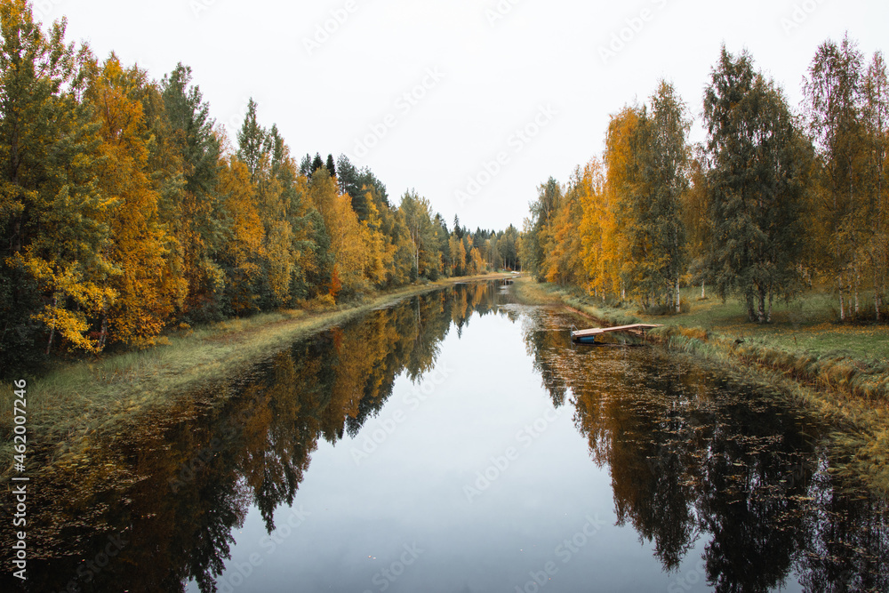 Fototapeta premium Autumn fairy tale in Kainuu, Finland. The colourful deciduous trees play with all their colours and reflect on the lake surface on a cloudy day. Orange, Green, blue colours