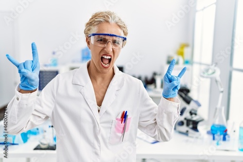 Middle age blonde woman working at scientist laboratory shouting with crazy expression doing rock symbol with hands up. music star. heavy concept.
