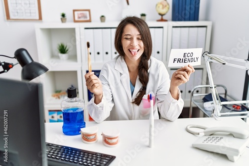 Young dentist woman holding eco friendly toothbrush sticking tongue out happy with funny expression.