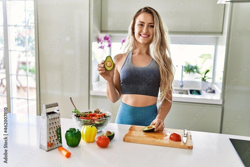 Young caucasian fitness woman wearing sportswear preparing healthy salad at the kitchen looking positive and happy standing and smiling with a confident smile showing teeth