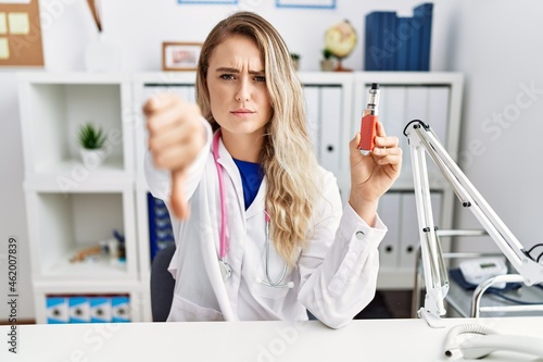 Young beautiful doctor woman holding electronic cigarette at the clinic with angry face, negative sign showing dislike with thumbs down, rejection concept