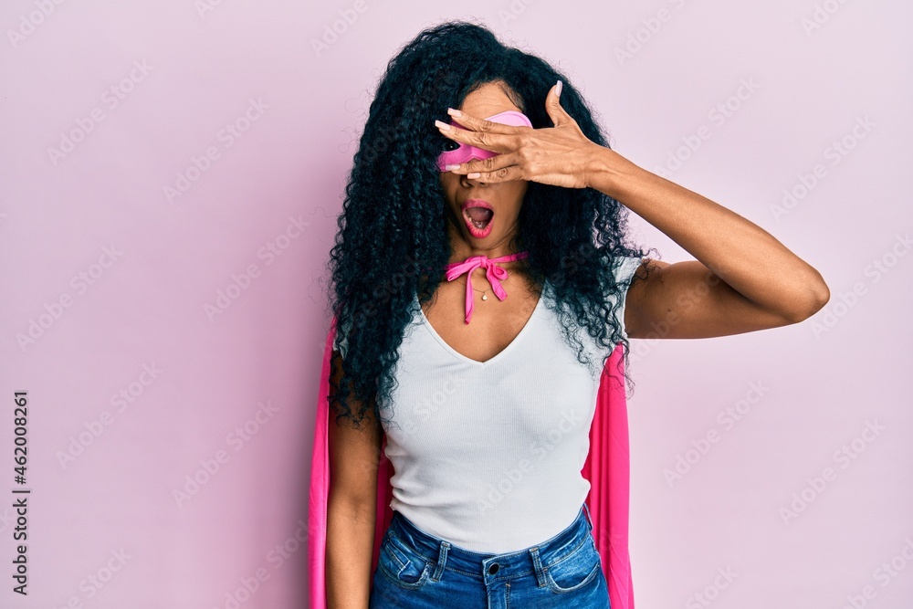 Middle age african american woman wearing super hero costume peeking in shock covering face and eyes with hand, looking through fingers with embarrassed expression. Middle age african american woman wearing super hero costume peeking in shock covering face and eyes with hand, looking through fingers with embarrassed expression.