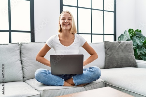 Young caucasian woman using laptop at home sitting on the sofa looking positive and happy standing and smiling with a confident smile showing teeth