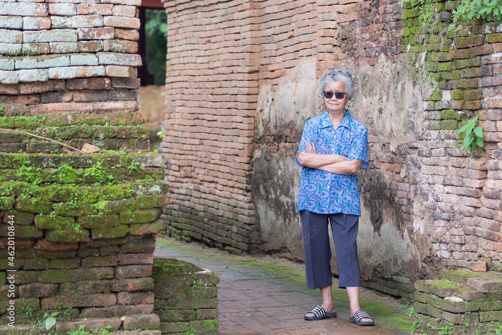 Portrait of an elderly Asian woman wearing sunglasses, arms crossed and looking at the camera with a smile while standing old brick wall background. Aged people and travel concept Portrait of an elderly Asian woman wearing sunglasses, arms crossed and looking at the camera with a smile while standing old brick wall background. Aged people and travel concept