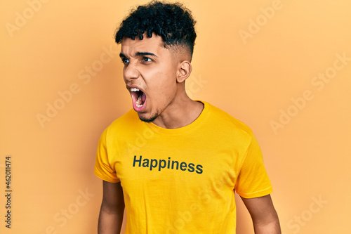 Young african american man wearing t shirt with happiness word message angry and mad screaming frustrated and furious, shouting with anger. rage and aggressive concept.