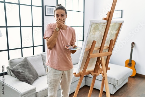Young hispanic man with beard painting on canvas at home laughing and embarrassed giggle covering mouth with hands, gossip and scandal concept