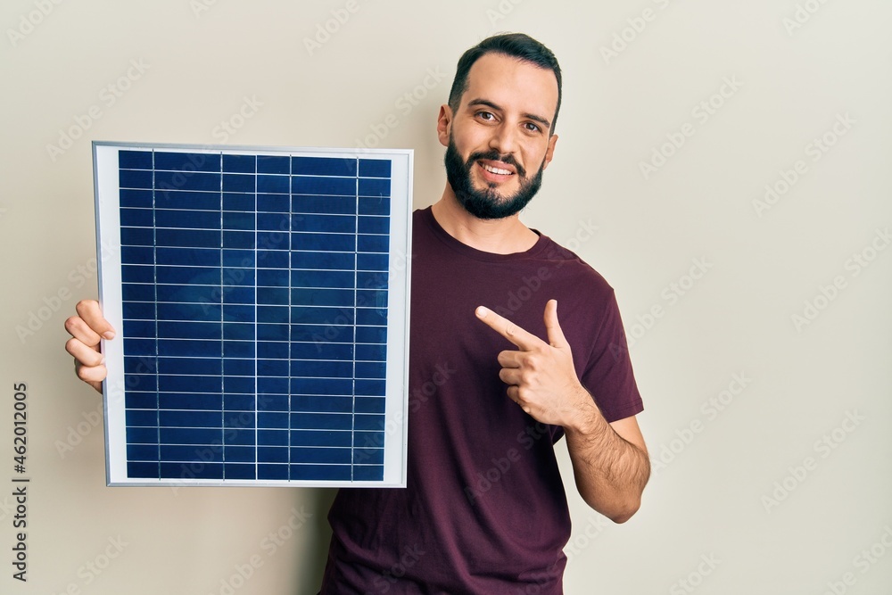 Young man with beard holding photovoltaic solar panel smiling happy pointing with hand and finger Young man with beard holding photovoltaic solar panel smiling happy pointing with hand and finger