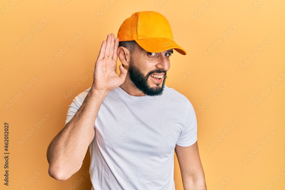 Young man with beard wearing yellow cap smiling with hand over ear listening and hearing to rumor or gossip. deafness concept.