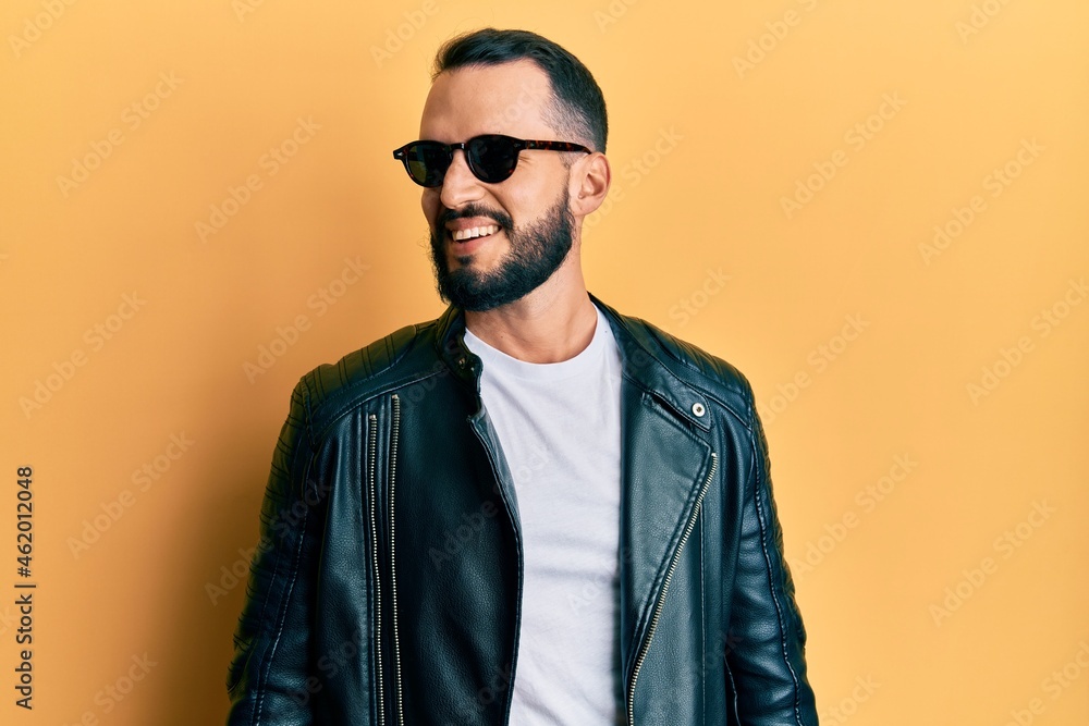 Fototapeta premium Young man with beard wearing black leather jacket and sunglasses looking away to side with smile on face, natural expression. laughing confident.