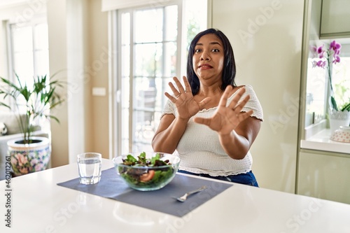 Young hispanic woman eating healthy salad at home afraid and terrified with fear expression stop gesture with hands  shouting in shock. panic concept.