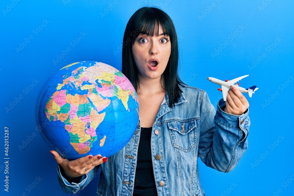 Fototapeta premium Young hispanic woman holding paper toy and world ball afraid and shocked with surprise and amazed expression, fear and excited face.