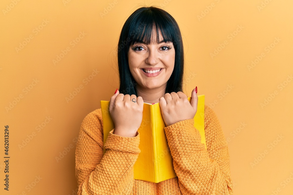 Young hispanic woman holding book smiling with a happy and cool smile on face. showing teeth. Young hispanic woman holding book smiling with a happy and cool smile on face. showing teeth.