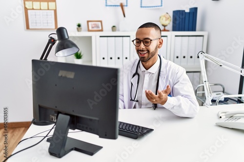 Young hispanic man wearing doctor uniform having video call at clinic