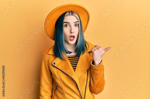Young modern girl wearing yellow hat and leather jacket surprised pointing with hand finger to the side, open mouth amazed expression.