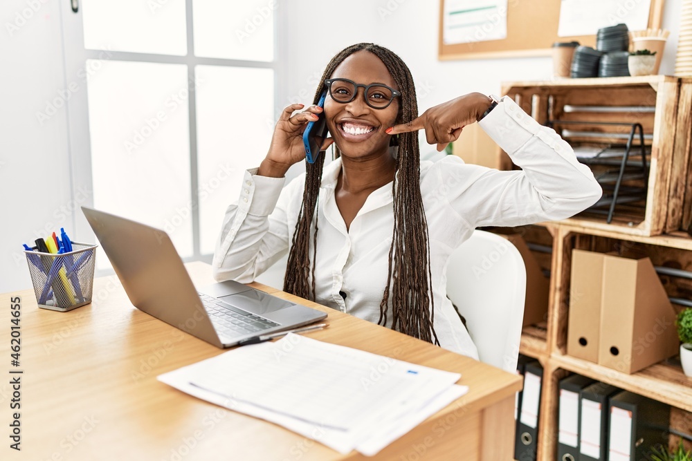 Black woman with braids working at the office speaking on the phone smiling cheerful showing and pointing with fingers teeth and mouth. dental health concept. Black woman with braids working at the office speaking on the phone smiling cheerful showing and pointing with fingers teeth and mouth. dental health concept.