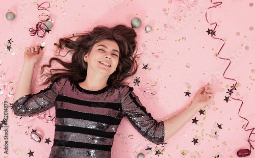 Cheerful girl lies on a pink background among confetti and serpentine, top view.