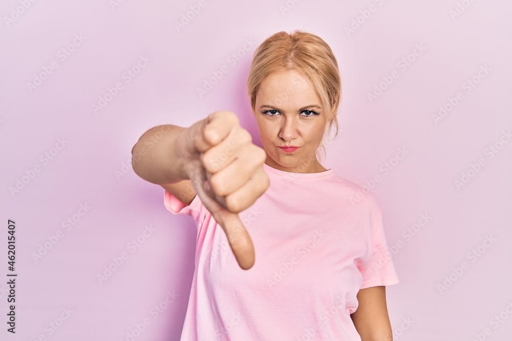 Young blonde woman wearing casual pink t shirt looking unhappy and angry showing rejection and negative with thumbs down gesture. bad expression. Young blonde woman wearing casual pink t shirt looking unhappy and angry showing rejection and negative with thumbs down gesture. bad expression.