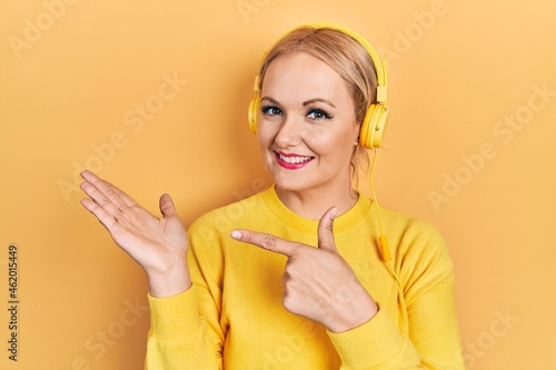 Young blonde woman listening to music using headphones amazed and smiling to the camera while presenting with hand and pointing with finger.