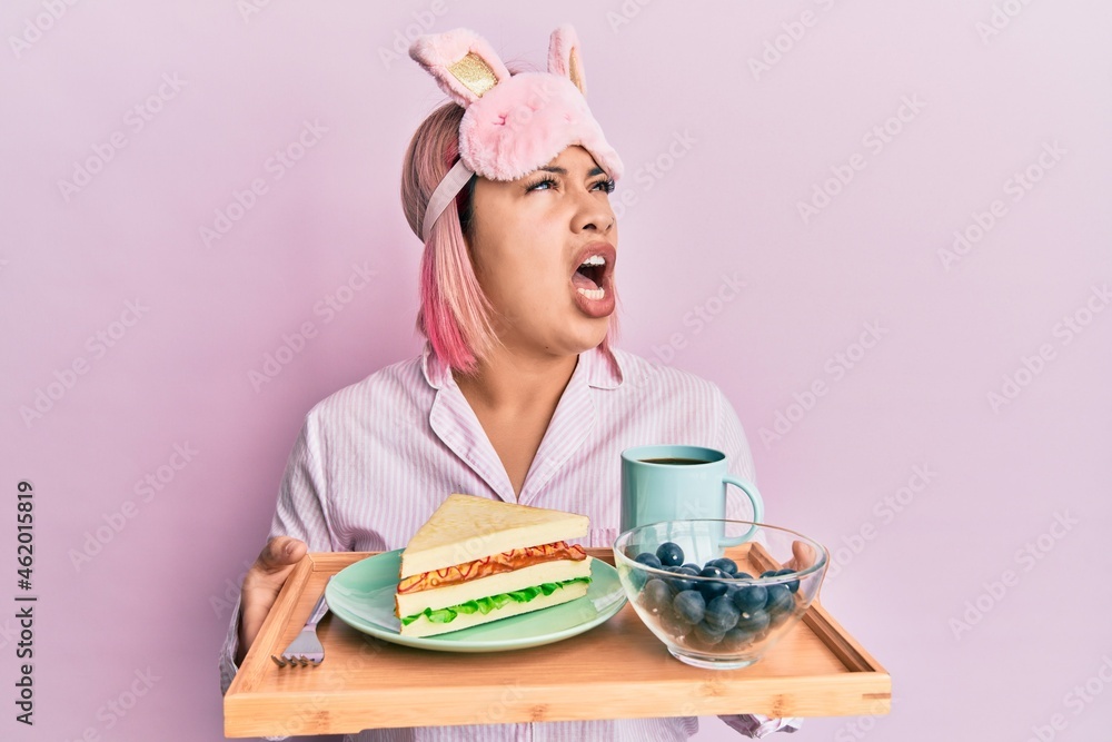 Hispanic woman with pink hair wearing pajama holding healthy breakfast angry and mad screaming frustrated and furious, shouting with anger looking up.