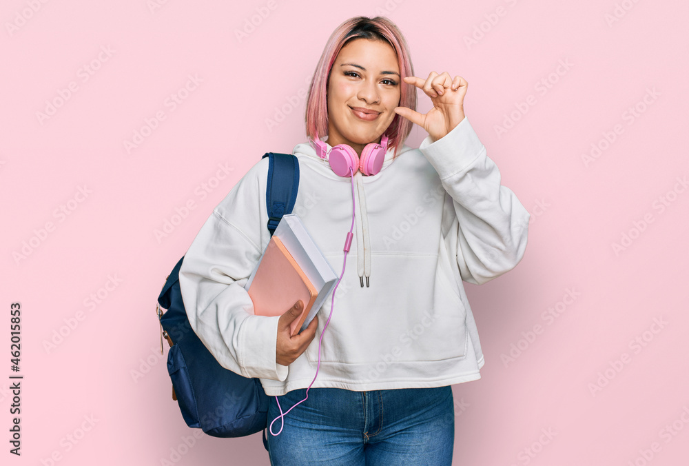 Hispanic woman with pink hair wearing student backpack and headphones smiling and confident gesturing with hand doing small size sign with fingers looking and the camera. measure concept. Hispanic woman with pink hair wearing student backpack and headphones smiling and confident gesturing with hand doing small size sign with fingers looking and the camera. measure concept.