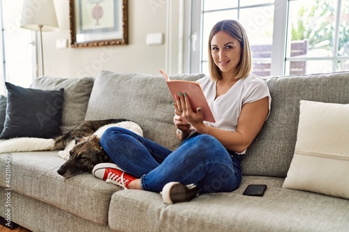 Young caucasian girl smiling happy sitting on the sofa with dog reading book at home.
