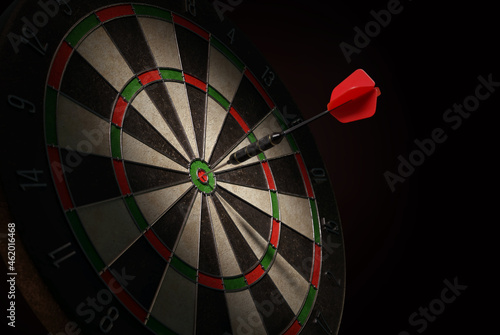 3D illustration of a shiny black dart with a red flight hitting the bulls-eye of a dartboard.