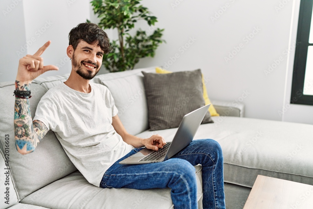 Hispanic man with beard sitting on the sofa smiling and confident gesturing with hand doing small size sign with fingers looking and the camera. measure concept.