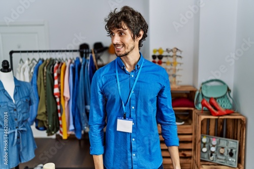Young hispanic man working as manager at retail boutique looking away to side with smile on face, natural expression. laughing confident.