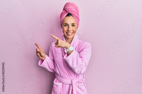 Young blonde woman wearing shower towel cap and bathrobe smiling and looking at the camera pointing with two hands and fingers to the side.