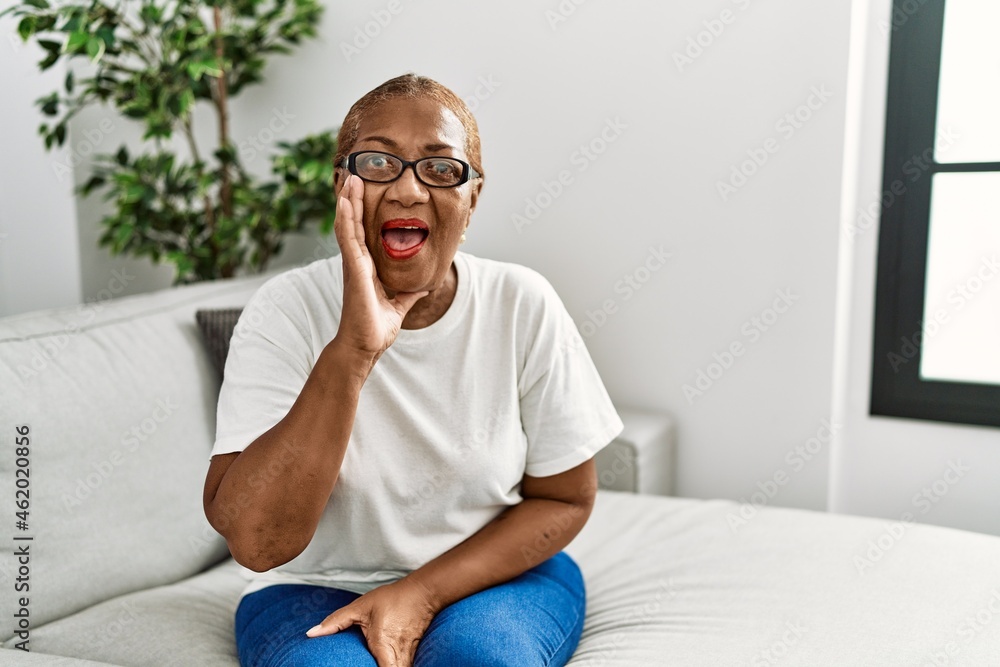 Mature hispanic woman sitting on the sofa at home shouting and screaming loud to side with hand on mouth. communication concept.