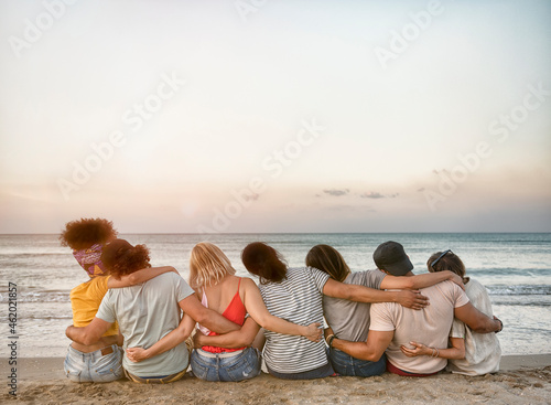 Rear view of sad multiracial group of teenage friends sitting on the sand of the beach looking at the horizon during sunset on a late summer day - Friendship Freedom Beach Summer Holiday Concept © charmedlightph