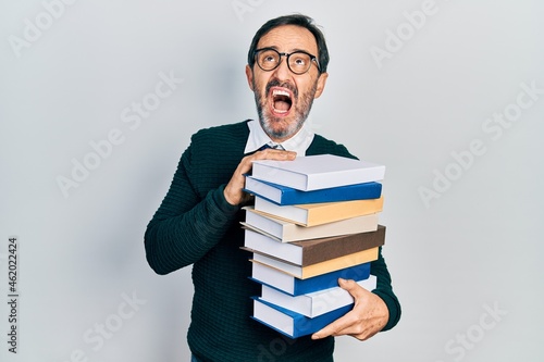 Middle age hispanic man holding a pile of books angry and mad screaming frustrated and furious, shouting with anger looking up.