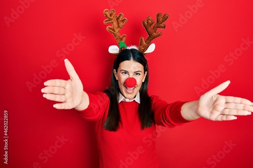 Young hispanic woman wearing deer christmas hat and red nose looking at the camera smiling with open arms for hug. cheerful expression embracing happiness.