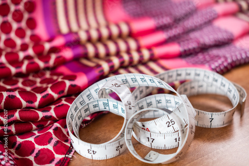 Close and selective focus of a tailor’s tape measure on a wooden table. In the background is some colourful fabric or material, intentionally out of focus